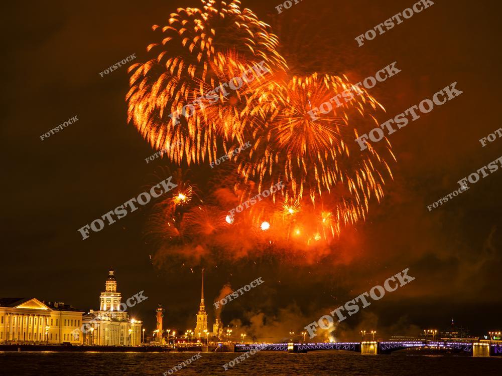 На фотографии изображён яркий фейерверк над Невой в Санкт-Петербурге. На переднем плане можно увидеть здание Кунсткамеры и подсвеченные Ростральные колонны. Освещённый Дворцовый мост добавляет сцене торжественности. Вечернее шоу подчёркивает красоту ночного города и его архитектурного наследия.